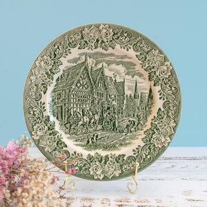 Può includere: Un piatto decorativo vintage verde e bianco con una scena di villaggio dettagliata. Il piatto ha un bordo floreale ed è esposto su un supporto dorato. Lo sfondo è azzurro.