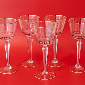 Può includere: Cinque calici da vino trasparenti con bordi dorati e dettagli decorativi rossi. I calici hanno gambi lunghi e poggiano su uno sfondo rosso. I calici sono eleganti e adatti per servire il vino.