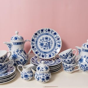 Puede incluir: Un juego de té completo de porcelana azul y blanca con diseños florales. El juego incluye tazas, platillos, platos, una crema, azucarero y dos teteras. La vajilla está dispuesta sobre una superficie blanca sobre un fondo rosa.