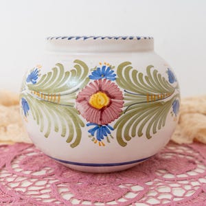 May include: A round ceramic vase with a white base featuring hand-painted floral motifs in shades of pink, yellow, blue, and green. The design includes a central pink flower with a yellow center, surrounded by green leafy patterns and blue accents.