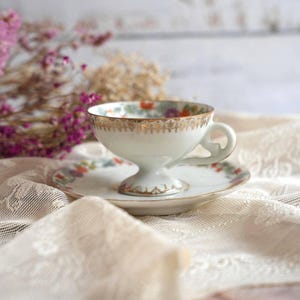 May include: A white porcelain teacup and saucer set featuring a delicate floral pattern in shades of orange, blue, and green. The rim of the cup and the edge of the saucer are decorated with a gold filigree design, and the cup has a pedestal base.