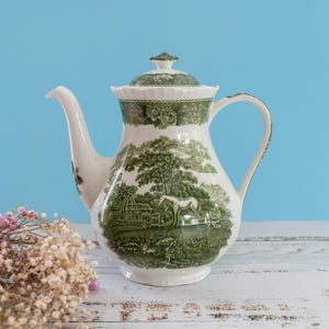 Può includere: Una teiera in ceramica bianca con un dettagliato motivo a trasferimento verde che raffigura un paesaggio pastorale con un cavallo, alberi e un edificio. La teiera è dotata di coperchio coordinato, manico curvo e un lungo beccuccio.