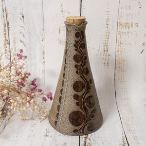 Può includere: Un vaso decorativo a forma di cono con un tappo di sughero. Il vaso è beige con un motivo marrone. Il motivo include elementi circolari e a spirale. Il vaso è alto circa 25 cm.