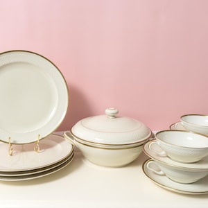 Pode incluir: Um conjunto de louça composto por pratos, uma terrina com tampa e chávenas de chá com pires na cor creme. Cada peça apresenta um padrão de diamante subtilmente em relevo ao longo da borda e um acabamento com um fino friso dourado.