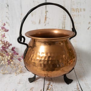 May include: A copper cauldron with a hammered texture, black handle and three small feet. The pot has a rounded shape and a shiny finish. The background is a rustic wooden surface.