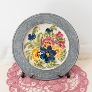 May include: A decorative plate with a grey, ornate rim. The centre features a floral design with yellow, blue, and red flowers on a cream background. The plate is displayed on a pink lace doily.