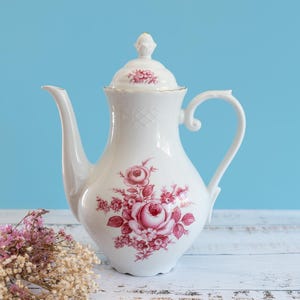 May include: A white porcelain teapot featuring a delicate pink floral rose pattern on the body and lid. The teapot has a curved handle, a long spout, and a removable lid with a finial. It is decorated with intricate embossed detailing.