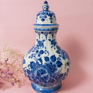 Op de afbeelding: Een hoge, wit en blauwe keramische vaas met een deksel. De vaas heeft ingewikkelde bloempatronen in blauwtinten. Het deksel is bekroond met een kleine bekroning. De vaas is een decoratief stuk, waarschijnlijk voor weergave of opslag.