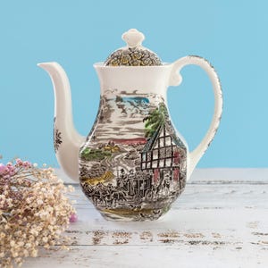 Może przedstawiać: Biały ceramiczny imbryk ozdobiony szczegółową czarno-białą grafiką przedstawiającą powóz konny i dom z muru pruskiego. Pokrywka posiada motyw kwiatowy, a uchwyt oraz dzióbek wykonane są z gładkiej, białej ceramiki bez dodatkowych zdobień.