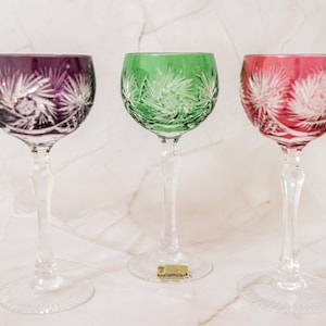 Könnte beinhalten: Drei Weingläser aus Kristall mit klaren Stielen und Füßen, mit tiefvioletten, smaragdgrünen und rubinroten Kuppen. Jede Kuppel ist mit aufwendigen, handgeschliffenen Stern- und Fächermustern verziert, die das klare Glas unter der Farbe zeigen.
