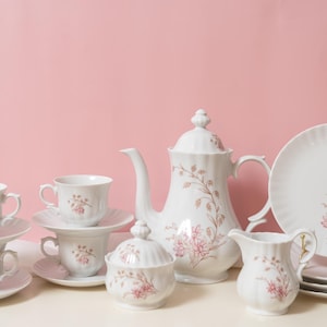 May include: A white porcelain tea set with pink floral patterns. The set includes teacups, saucers, a teapot, a sugar bowl, a creamer, and plates. The tea set is arranged on a white surface against a pink backdrop.