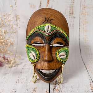 May include: A wooden mask with a carved face, decorated with green and yellow beaded details, seashells, and gold-coloured earrings. The mask has a smiling mouth and is likely a decorative item.