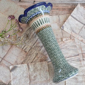 Könnte beinhalten: Eine hohe Keramikvase mit einem ausgestellten, gewellten Rand mit dunkelblauer Kante. Der Körper ist mit einem sich wiederholenden Muster aus grünen Ovalen verziert, während der Hals ein Band aus kleinen grünen Blattmotiven und orangefarbenen Punkten zeigt.