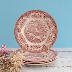 Può includere: Una pila di tre piatti rotondi in ceramica con un motivo a trasferimento rosa e bianco. Il centro di ogni piatto raffigura un paesaggio con un edificio e una persona a cavallo, circondato da un bordo floreale dettagliato.
