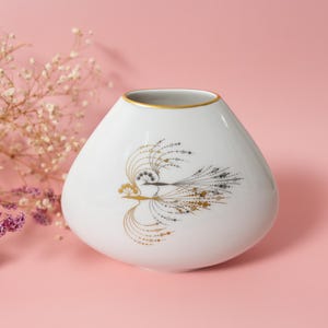 May include: White ceramic vase with a gold rim and a stylized bird design in gold and black. The vase has a rounded shape and sits on a pink background. Dried flowers are in the background.