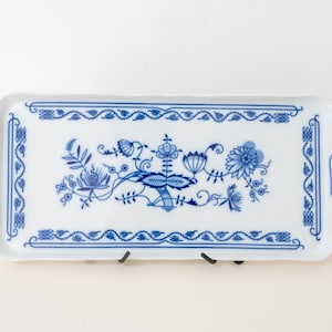 May include: White ceramic serving tray with a blue floral pattern. The rectangular tray features a decorative border and handles. The blue design includes flowers and leaves. The tray is supported by a black metal stand. The tray's dimensions are in centimetres.