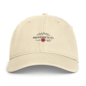 May include: A cream-colored baseball cap featuring a central graphic with the text PROVERBS 31 CO. in black, surrounded by a delicate black leaf vine design and a small pink circular accent positioned directly below the text.
