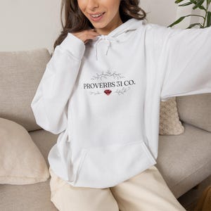 May include: A white long-sleeve hooded sweatshirt featuring a front kangaroo pocket and a drawstring hood. The chest displays the black text PROVERBS 31 CO. centered between two delicate leafy vine illustrations and a small red diamond graphic.