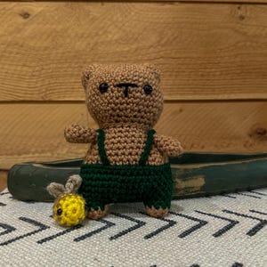 May include: A small, light brown crocheted bear toy wearing dark green overalls with straps. The bear has black bead eyes and a stitched nose. Beside the bear sits a tiny, round, yellow and black crocheted bee with small wings.