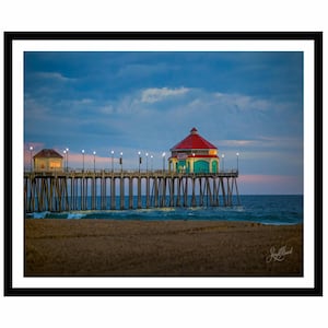 Puede incluir: Fotografía escénica de un muelle que se extiende hacia el océano al atardecer. El muelle presenta un edificio con techo rojo y está iluminado por farolas. El cielo es una mezcla de tonos azules y rosados, con una playa arenosa en primer plano.