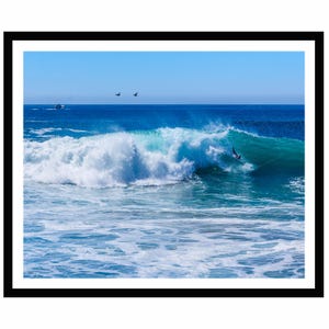 May include: A framed photograph of a large turquoise ocean wave breaking with white sea foam. A surfer in a black wetsuit is riding the wave. Two birds fly in the clear blue sky above the horizon, and a small boat is visible in the distance.