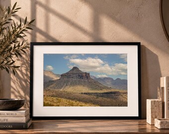 May include: A framed landscape photograph featuring a prominent, dark, cone-shaped mountain peak rising above a dry, golden-brown desert terrain under a bright blue sky with scattered white clouds, presented in a simple black frame with a white mat.