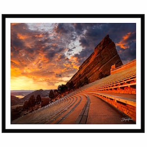 May include: A framed landscape photograph of Red Rocks Amphitheatre at sunset. The image captures the natural rock formations and tiered seating under a vibrant sky of orange, yellow, and blue hues. The photograph is set in a black frame.