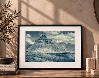 May include: A framed landscape photograph featuring a prominent, craggy mountain peak under a cloudy sky. The image is rendered in a monochromatic blue-toned palette, displayed within a black frame with a white mat, resting on a wooden surface.