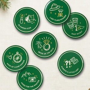 Puede incluir: Un conjunto de siete chapas circulares verdes con ilustraciones de líneas blancas y texto. Los diseños incluyen temas como una brújula, un anillo de diamantes, una cámara y equipo de acampada, con frases como Glamper Icon.