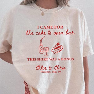 Op de afbeelding: Ivoor t-shirt met rode tekst en afbeeldingen. De tekst op het shirt luidt "I came for the cake & open bar" met een fles, glas en taart illustratie. Er staat ook "This shirt was a bonus" en de namen "Chloe & Chris" met "Phoenix, May 30".
