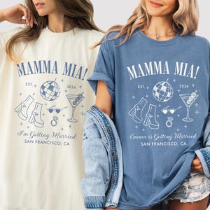 Puede incluir: Dos camisetas de manga corta en color crema y azul con el texto MAMMA MIA! EST. 2026, I'm Getting Married o Emma is Getting Married, y SAN FRANCISCO, CA. Los gráficos incluyen una bola de discoteca, botas de plataforma, gafas de corazón y un martini.