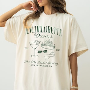 May include: Ivory-colored t-shirt with a moss green graphic that reads "BACHELORETTE Diaries" with a cocktail, crown, sunglasses, and crossed utensils. The text "Me? The Bride? Shut up!" and "SAN FRANCISCO, CA" are also printed on the shirt.