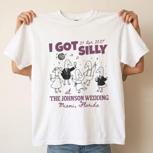 Op de afbeelding: Een wit t-shirt met een afbeelding van vijf cartoon-ganzen in formele trouwkleding, inclusief pakken en jurken. De tekst luidt I GOT SILLY 24 Apr 2027 AT THE JOHNSON WEDDING Miami, Florida, vergezeld door een discobal en muzieknoten.