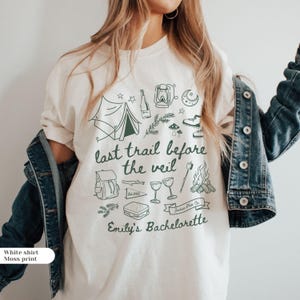 May include: Cream-coloured t-shirt featuring a moss green graphic with camping-themed illustrations and the text "last trail before the veil" and "Emily's Bachelorette". A denim jacket is worn over the shirt.