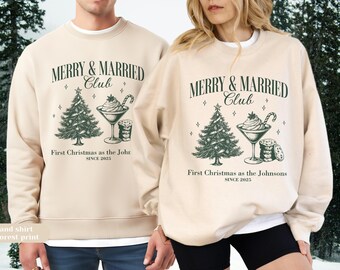 Può includere: Due felpe girocollo color sabbia con una stampa grafica verde scuro. Il design include un albero di Natale, un bicchiere da cocktail con panna montata, un bastoncino di zucchero e biscotti, con il testo MERRY & MARRIED Club, First Christmas as the Johnsons e SINCE 2025.
