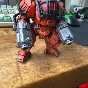 Może przedstawiać: Szczegółowa, pomarańczowo-srebrna miniaturowa figurka robota. Robot ma głowę w kształcie klatki, masywne ramiona i nogi i jest ustawiony na drewnianej powierzchni. Figurka wydaje się być modelem lub zabawką, prawdopodobnie do gry stołowej lub ekspozycji.