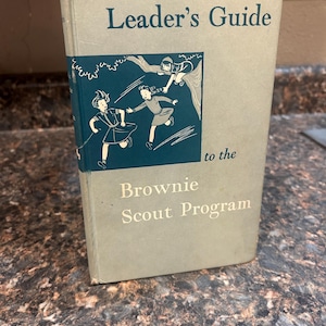 Może przedstawiać: Vintage książka "Leader's Guide to the Brownie Scout Program". Okładka jest jasno-szaro-niebieska z tytułem w ciemnoniebieskim kolorze. Kolorowa ilustracja harcerek Brownie znajduje się z przodu. Książka jest lekko zużyta.