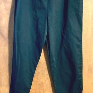 May include: A pair of solid dark teal trousers featuring a high-waisted design with a central zipper fly and button closure. The pants have a straight-leg cut with a clean, minimalist appearance and a smooth, matte fabric finish throughout the garment.