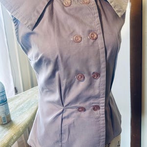 Puede incluir: Top sin mangas de color lavanda con diseño cruzado. El top presenta un cuello ancho y seis botones a juego. La tela parece ser un material liso y tejido. La prenda se exhibe en un maniquí.