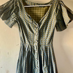May include: A dress featuring a green and blue plaid pattern with a V-neckline, short pleated sleeves, and a front button-down closure. The bodice is fitted, transitioning into a pleated skirt, and the garment is displayed on a white hanger.