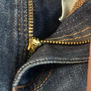 以下が含まれることがあります： デニムジーンズのジッパーのクローズアップ。ジッパーは金色で部分的に開いており、歯と生地の質感が現れています。デニムは濃い青色で、ステッチが見えます。