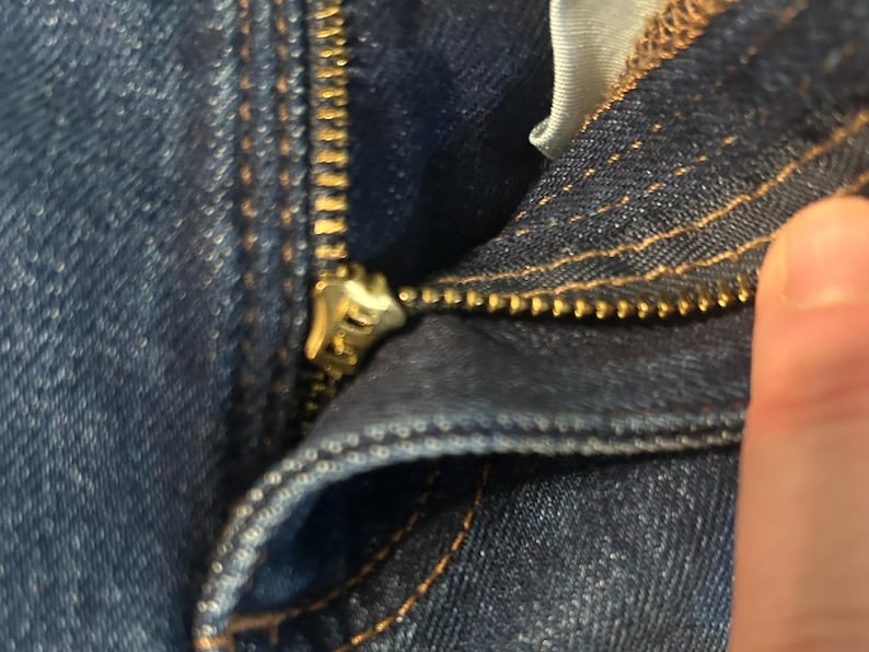 以下が含まれることがあります： デニムジーンズのジッパーのクローズアップ。ジッパーは金色で部分的に開いており、歯と生地の質感が現れています。デニムは濃い青色で、ステッチが見えます。