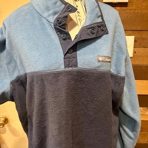 Puede incluir: Un jersey de forro polar azul con canesú y cuello azul marino. El jersey tiene cierre de botones y un pequeño logotipo de Columbia en el pecho. La prenda está hecha de un material suave y texturizado.