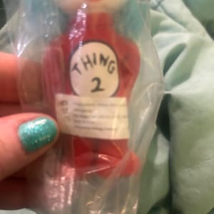 May include: A red plush toy of "Thing 2" from Dr. Seuss's "The Cat in the Hat" series, packaged in clear plastic. The toy has blue hair, a white face, and a red outfit with the number "2" on the front.