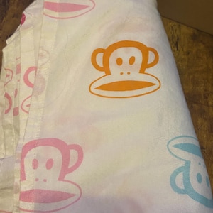 Puede incluir: Una sábana de tela blanca con un patrón repetitivo de caras de mono estilizadas en varios colores, incluyendo naranja, rosa y azul claro. Las caras de mono son dibujos lineales simples con orejas grandes y bocas ovaladas impresas en la superficie.