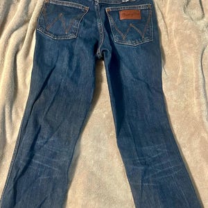 以下が含まれることがあります： バックポケットにWranglerのロゴが入った濃いブルーデニムジーンズ。ジーンズは、茶色の革パッチが付いたクラシックな5ポケットスタイルです。ジーンズは丈夫なデニム生地で作られています。