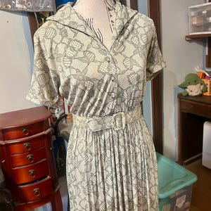Puede incluir: Vestido vintage crema con un estampado floral de encaje gris. Tiene mangas cortas, cuello camisero y cinturón a juego con hebilla rectangular. La falda es plisada.