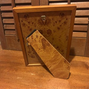 May include: Wooden picture frame with a burl wood veneer back and stand. The frame has a light brown wooden border. The back of the frame has a decorative burl wood pattern. The stand is also made of burl wood.