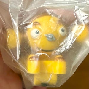 Könnte beinhalten: Eine gelbe Spielzeugfigur mit großen Augen, einem kleinen Nasenring und einem lächelnden Mund, verpackt in einer durchsichtigen Plastiktüte. Die Figur hat einen runden Kopf und Arme sowie einen rechteckigen Körper. Das Spielzeug hat eine leuchtend gelbe Farbe.