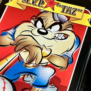 May include: A rectangular metal tin featuring a graphic of the cartoon character Taz dressed in a blue and white baseball uniform. The character holds a baseball bat, with a baseball in the foreground. A yellow banner reads M.V.P. TAZ.